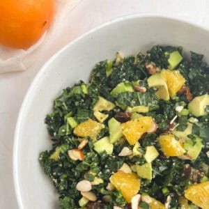 partial overhead view of white bowl containing kale citrus salad with an orange to the upper left