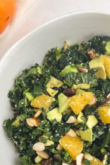 partial overhead view of white bowl containing kale citrus salad with an orange to the upper left