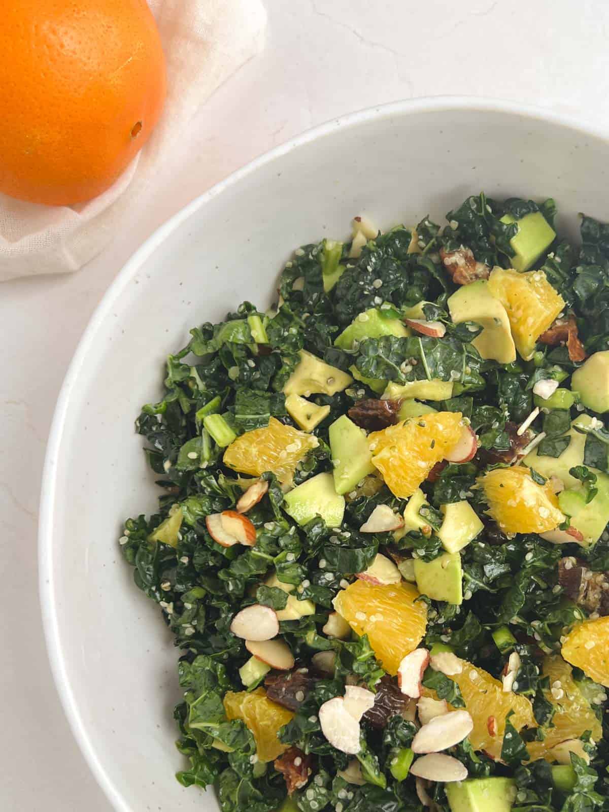 partial overhead view of kale citrus salad in a white bowl next to an orange in the top left corner