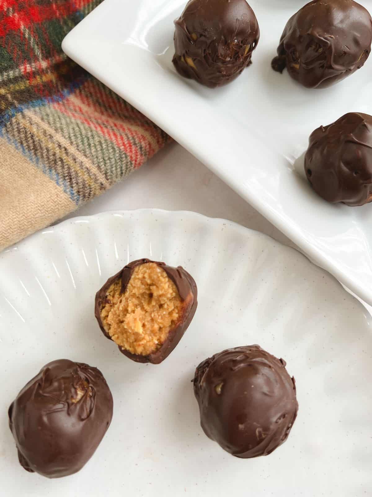 3 no bake chocolate peanut butter balls on a plate with a bite out of one, with rectangular serving tray and plaid cloth in the background