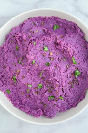 overhead view of a white bowl containing purple mashed potatoes on a marble background