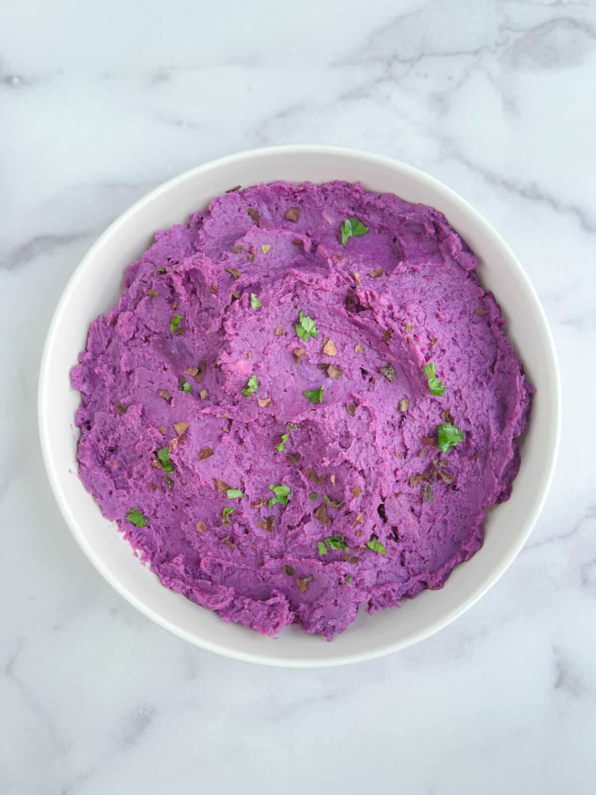 overhead view of a white bowl of purple mashed potatoes garnished with dulse flakes and parsley leaves on a marble background