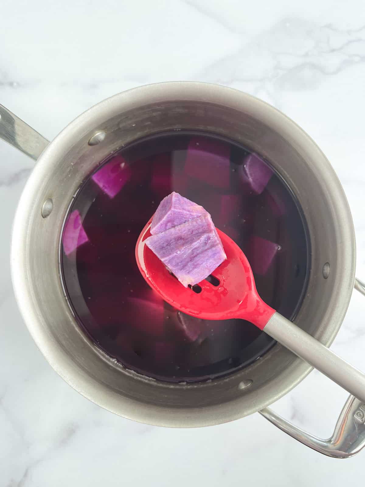 overhead view of stainless steel pot containing diced purple potatoes in water with a red silicone spoon overhead holding a couple potato pieces