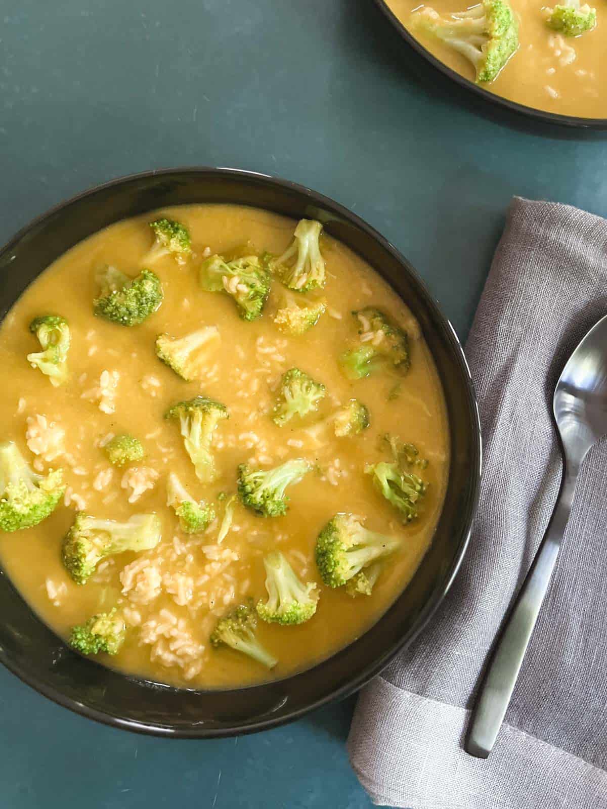 black bowl of vegan broccoli cheddar soup on a blue-green background next to a gray napkin and spoon