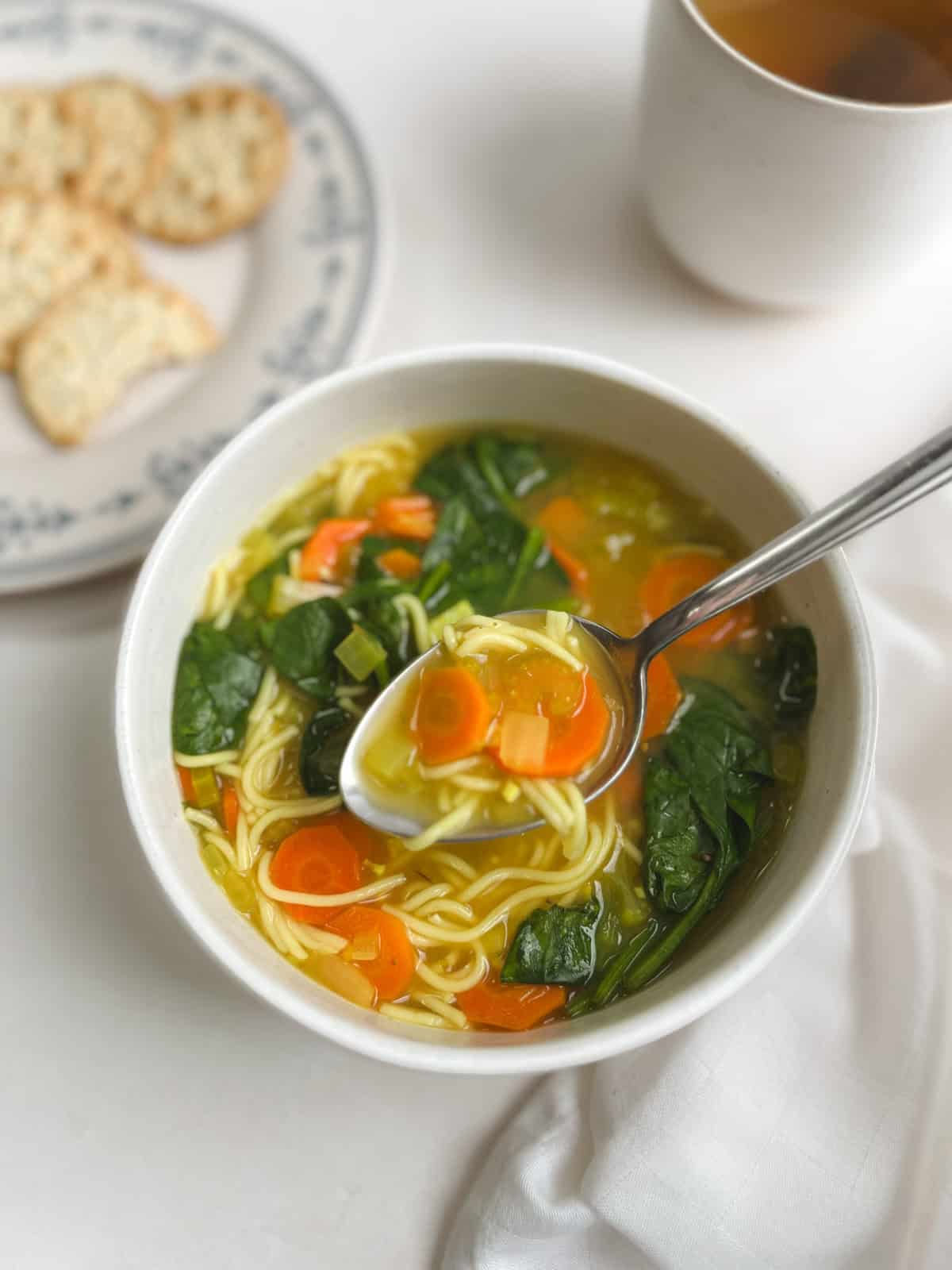 closeup of a spoonful of immunity soup being lifted out of a white bowl with a plate of crackers and a cup of tea in the background