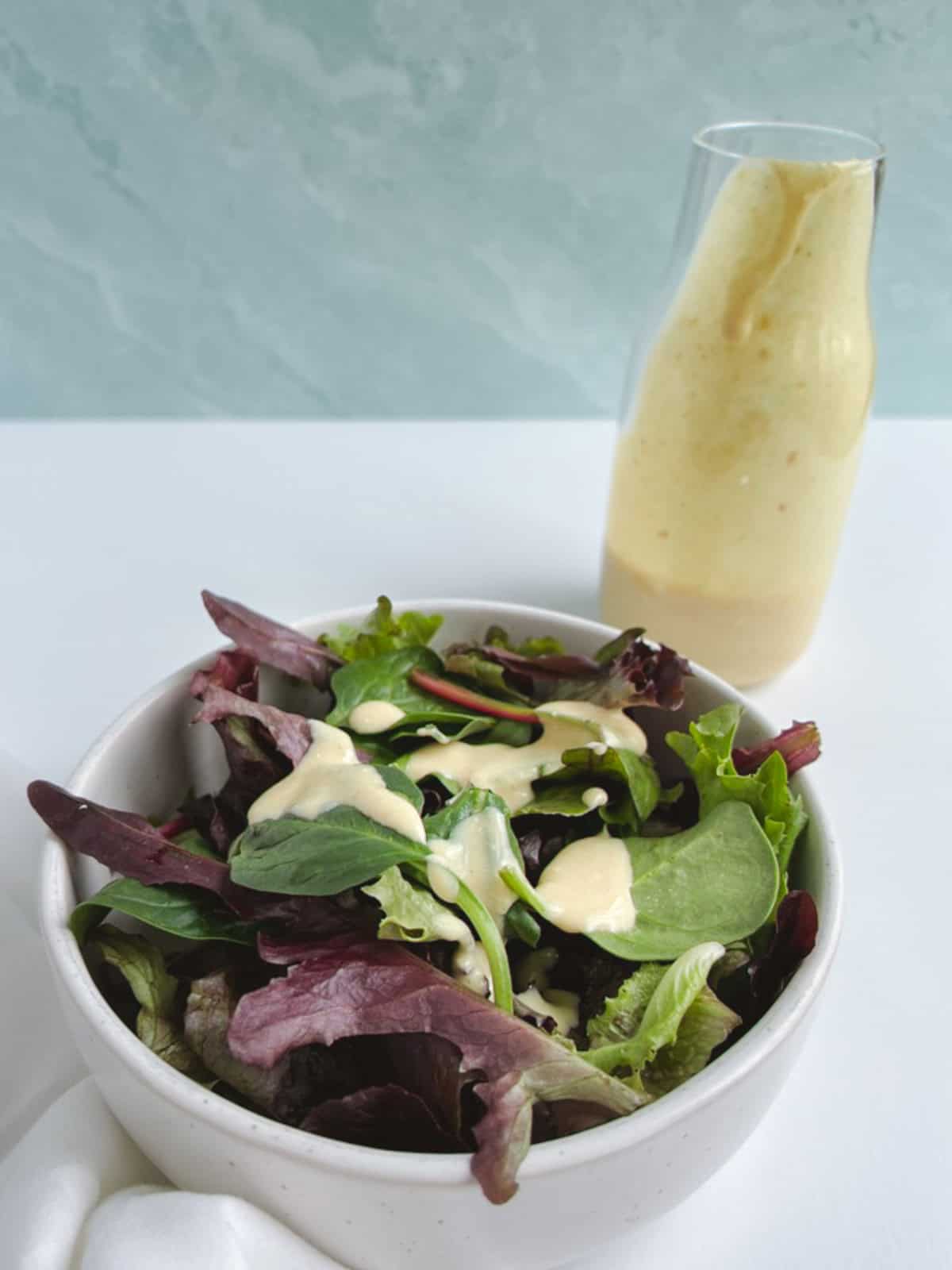 small white bowl of salad topped with vegan caesar dressing with glass bottle of dressing in the background