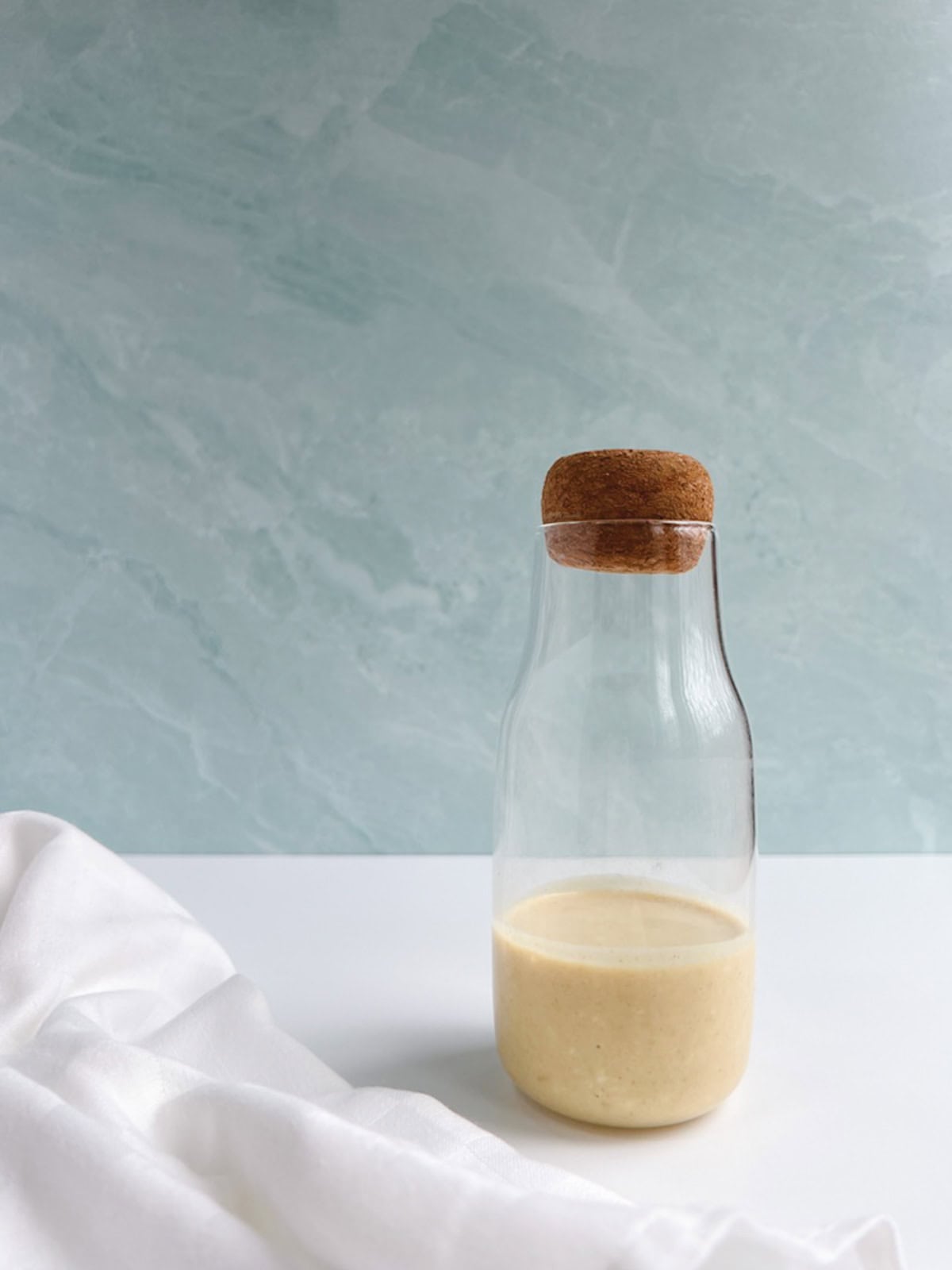 glass bottle of vegan caesar dressing on a white table in front of a blue-green background