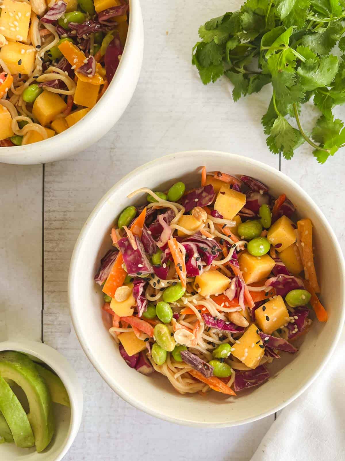 bowl of mango noodle salad with peanut dressing next to sliced avocado and cilantro