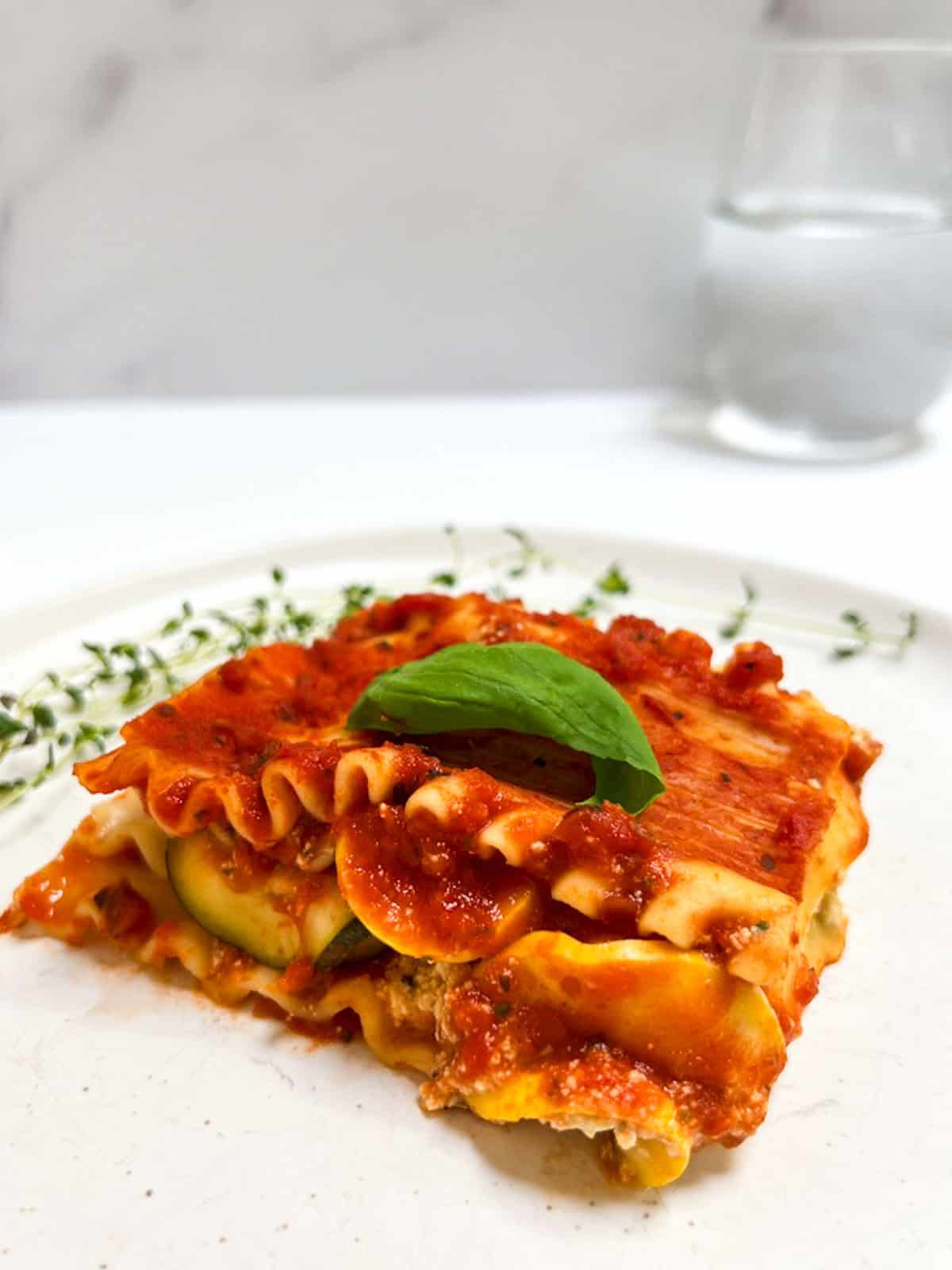 A slice of vegan zucchini lasagna on a plate with a glass of water