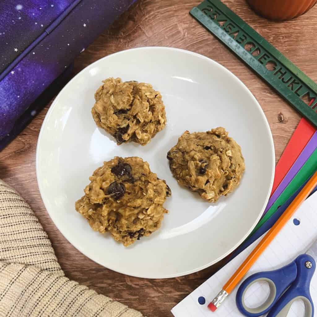 Three breakfast cookies sit on a white plate next to a backpack and pencils.