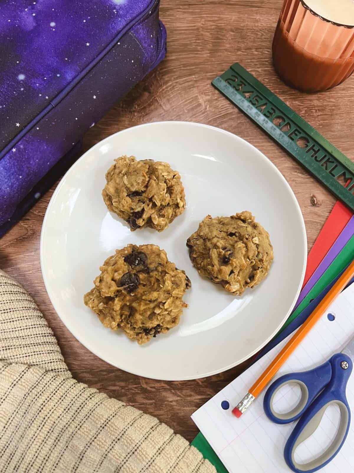 Three oatmeal breakfast cookies sit on a plate next to a backpack and pencils.