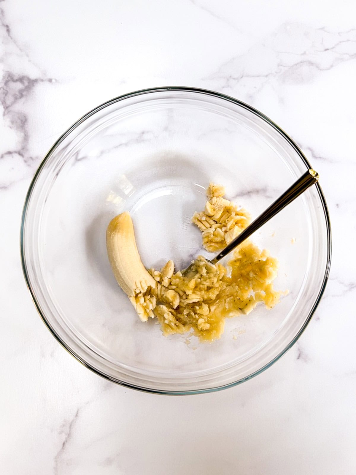 A bowl with a partially mashed banana and fork on a marble counter.